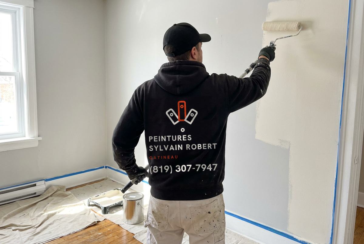 Peintre vu de dos, en chandail noir à capuchon aux couleurs de l’entreprise, qui roule un mur dans une chambre, toiles de protection au sol